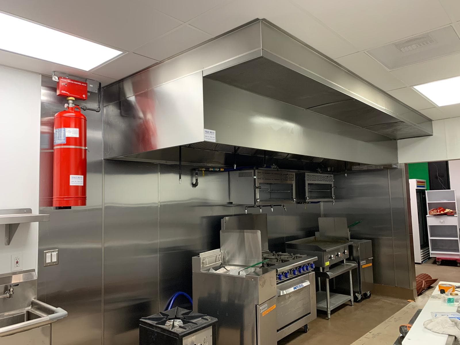 A commercial kitchen hood with its fire suppression tank mounted beside it, the ceiling still open